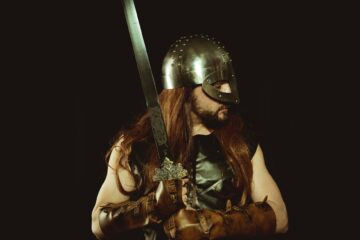 long haired man in metal helmet and leather gloves holding silver sword